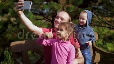 一个带孩子的男人坐在长凳上的智能手机上拍照。 幸福的一家人在公园里自拍。 家庭